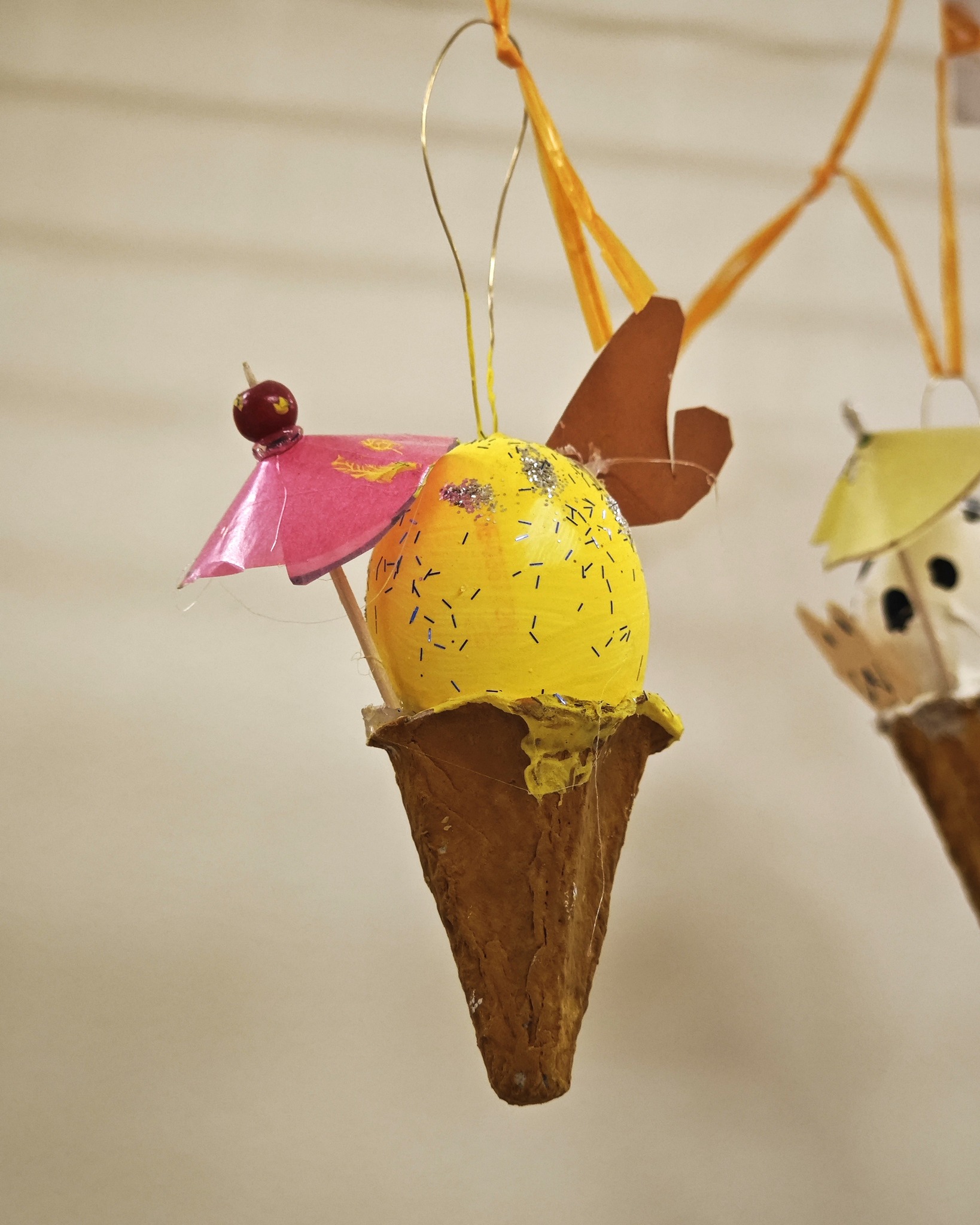 Dieser Alternativtext wurde KI-generiert: Eine dekorative, selbstgemachte Figur in Form eines Eises hängt an einer Schnur. Das Eis besteht aus bunten Materialien und hat eine Kirsche als Garnierung.