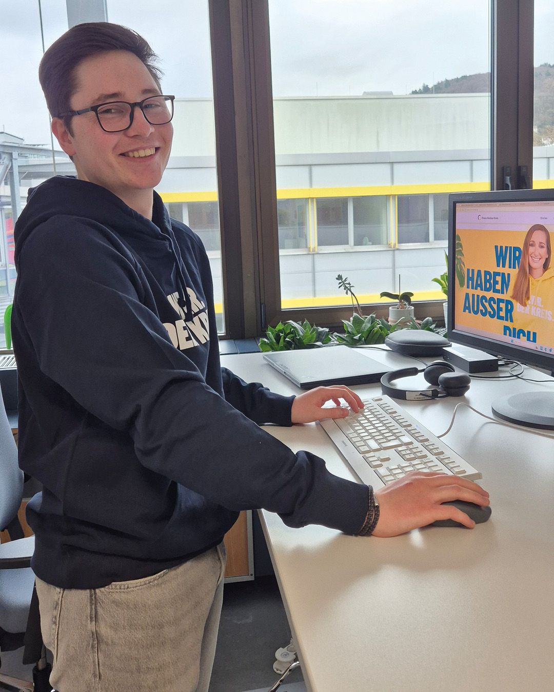 Dieser Alternativtext wurde KI-generiert: Eine Person sitzt an einem Schreibtisch vor einem Computer und lächelt. Im Hintergrund ist durch ein Fenster eine Landschaft zu sehen.