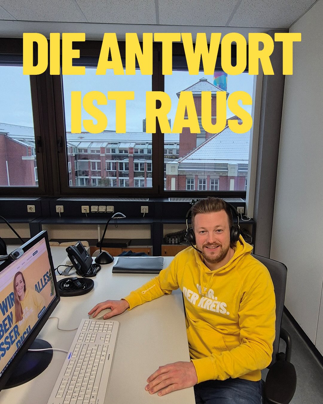 Dieser Alternativtext wurde KI-generiert: Ein Mensch sitzt vor einem Schreibtisch mit einem Computer, einem Mikrofon und anderen Geräten. Im Hintergrund ist eine Stadtansicht durch ein Fenster zu sehen.