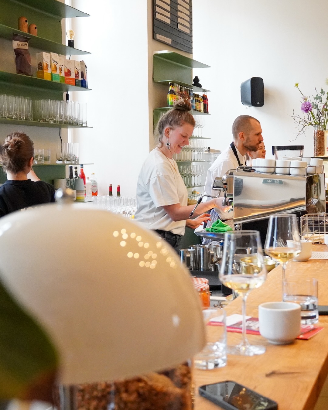 Dieser Alternativtext wurde KI-generiert: Ein Innenraum mit einer Theke und Regalen im Hintergrund. Mehrere Personen sind zu sehen, einige scheinen in einem Café zu arbeiten und Getränke zuzubereiten.