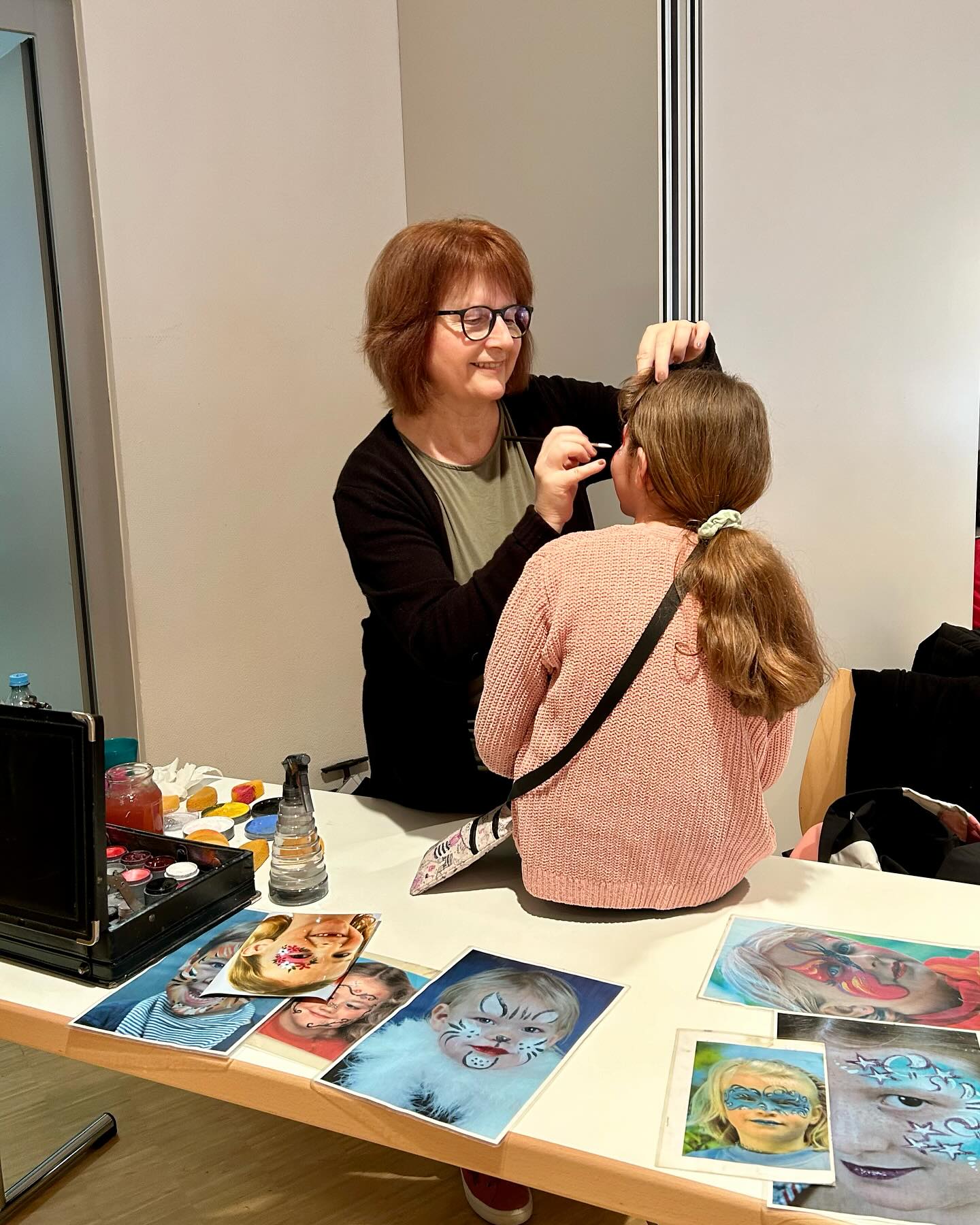Eine Person schminkt eine andere Person am Tisch. Auf dem Tisch liegen Fotos von geschminkten Gesichtern und Schminkutensilien.
