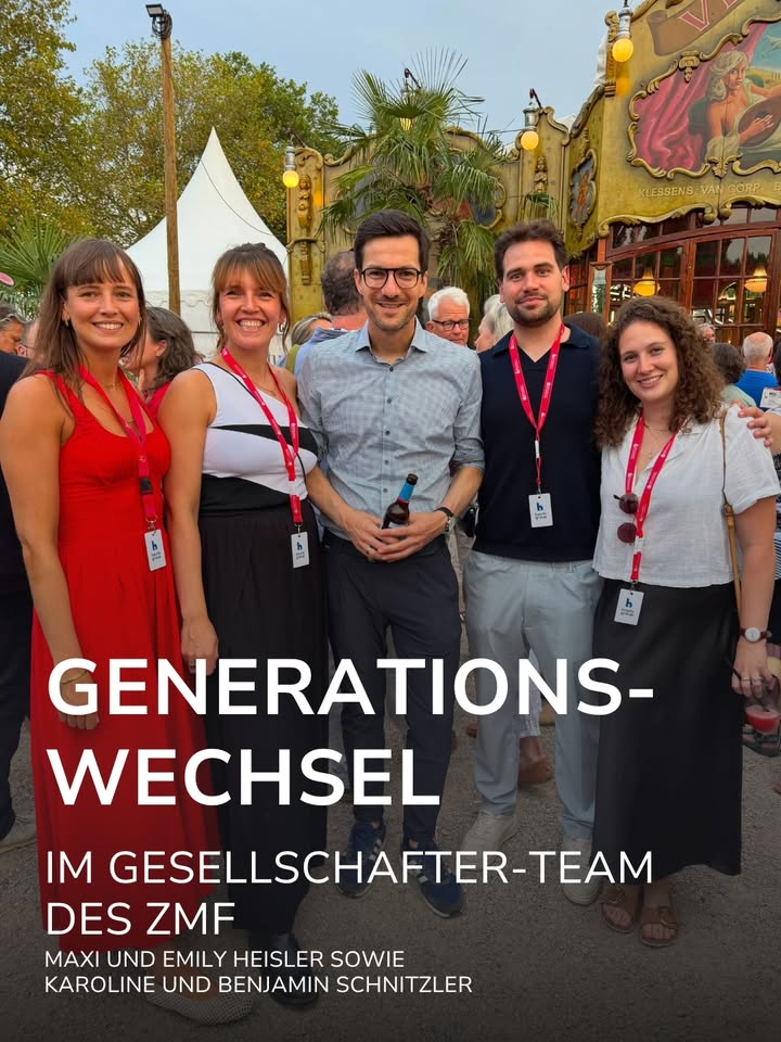 Fünf Personen stehen im Freien vor einem Karussell und lächeln, wobei sie rote Lanyards tragen. Ein großer Textüberzug lautet „GENERATIONSWECHSEL IM GESELLSCHAFTER-TEAM DES ZMF“, gefolgt von einer Auflistung von vier Namen. (KI-Alternativtext)
