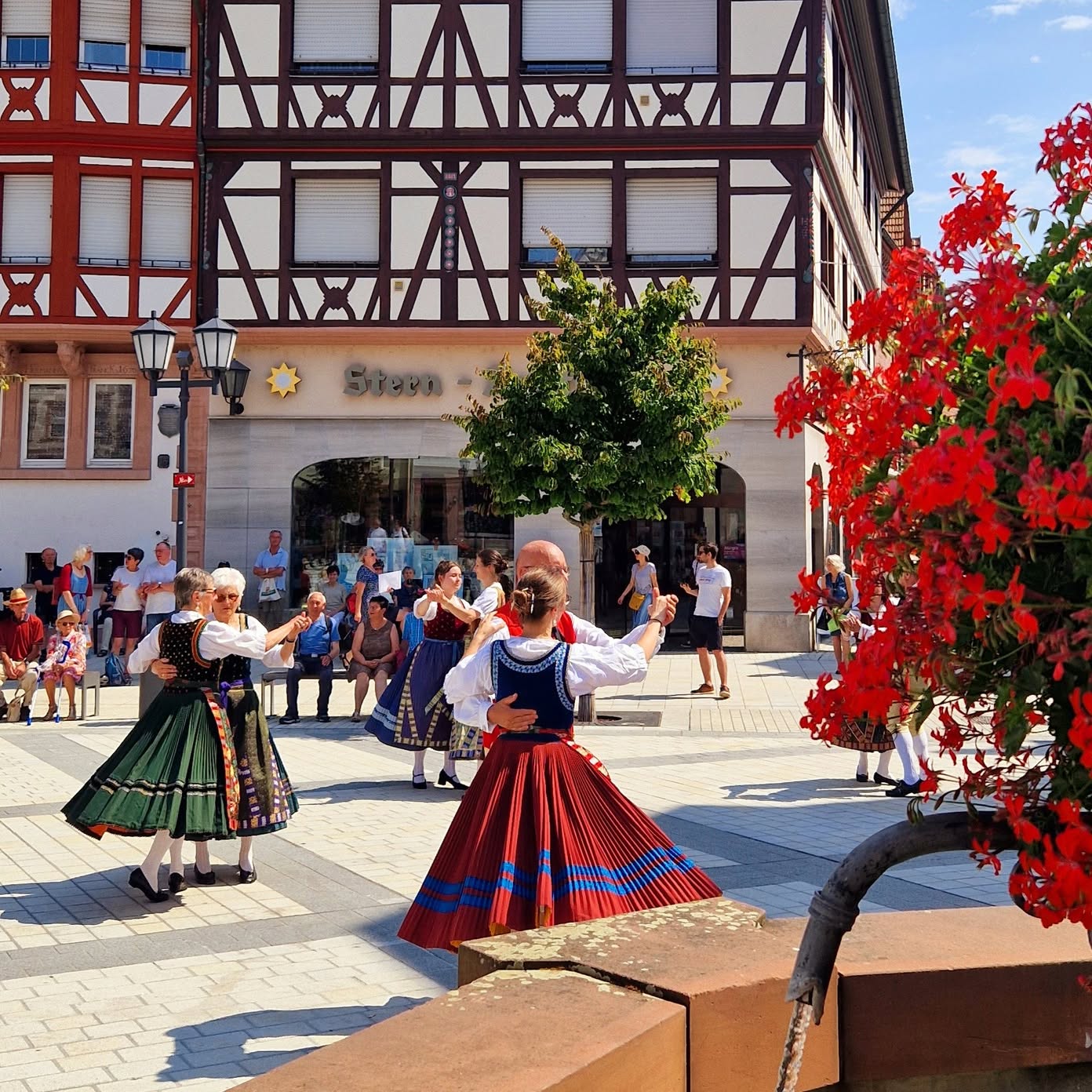 Auf einem Platz tanzen mehrere Personen in Tracht. Im Hintergrund ist ein Fachwerkhaus zu sehen.