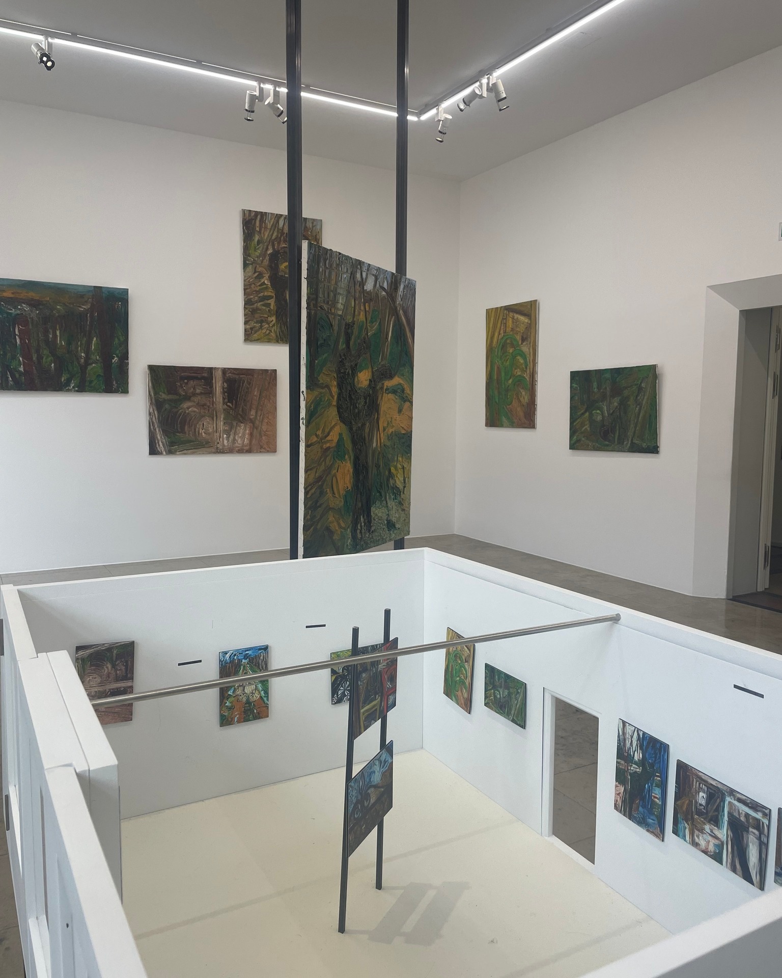 Dieser Alternativtext wurde KI-generiert: Ein heller Raum mit weißen Wänden, in dem mehrere farbige Gemälde hängen. Die Kunst ist auf mehreren Ebenen ausgestellt, die durch eine Balustrade getrennt sind.