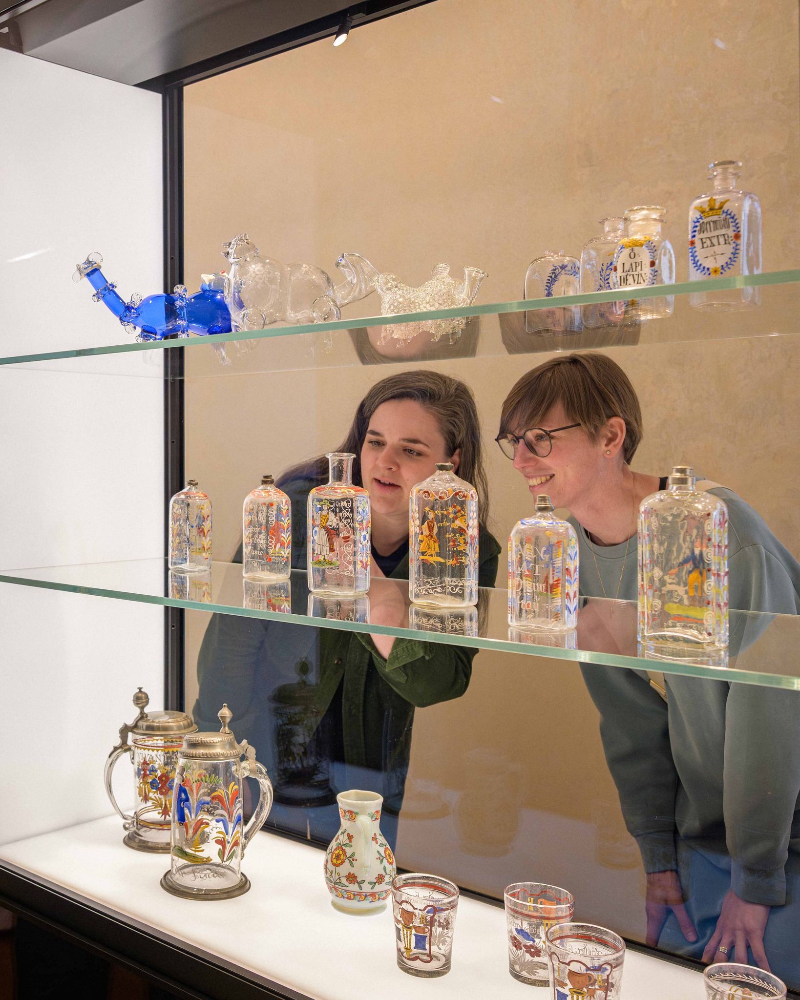 Zwei Personen betrachten eine Vitrine mit historischen Glasobjekten. Die Regale zeigen Glasfiguren von Kamelen, beschriftete und bemalte Flaschen sowie Krüge und Becher mit bunten Motiven. (KI-Alternativtext)