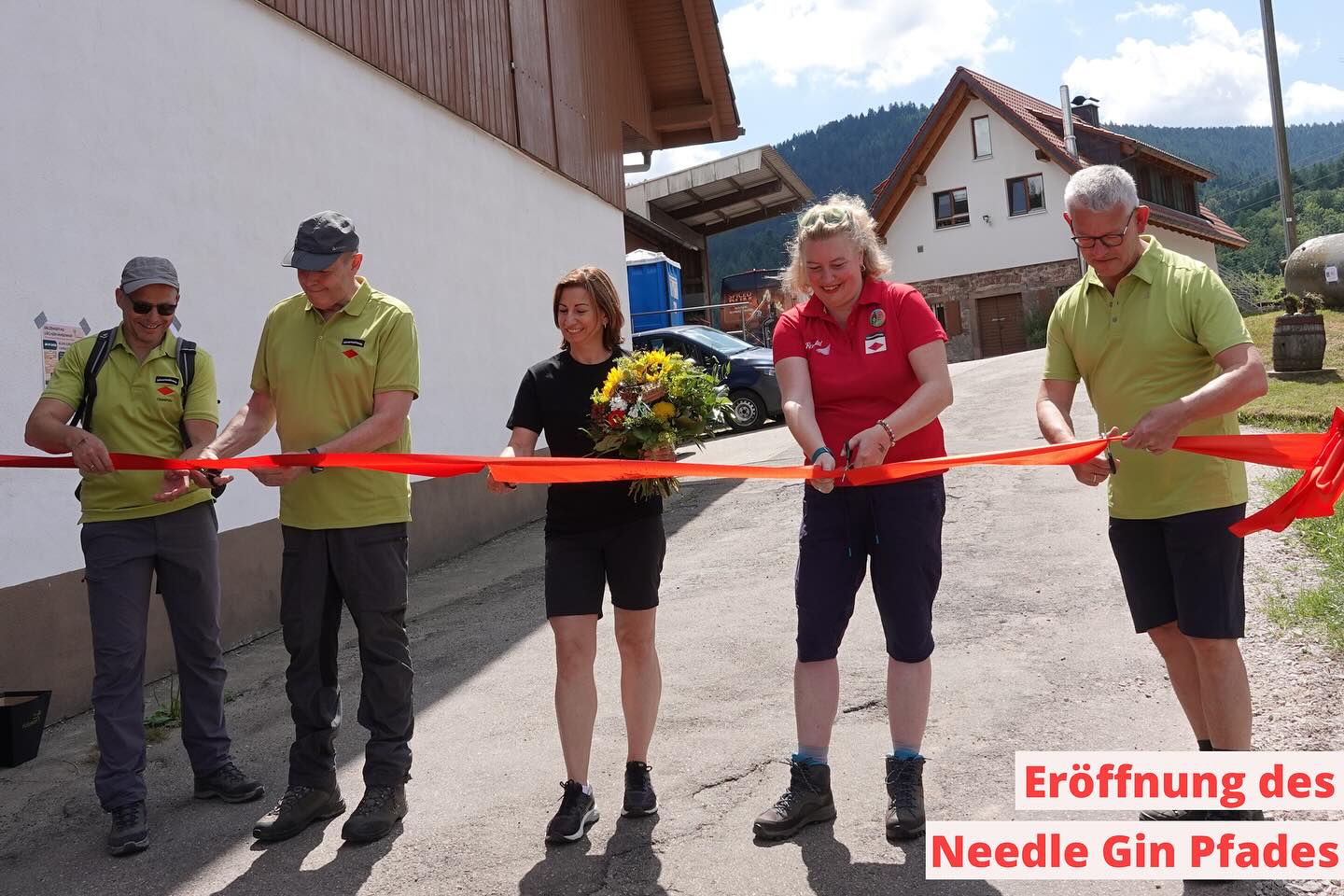 mehrere Personen durchschneiden ein rotes Band im Freien, wahrscheinlich bei einer Eröffnungszeremonie. Im Hintergrund sind ein Gebäude und Berge zu sehen.