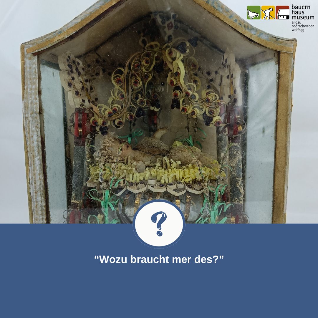 Dieser Alternativtext wurde KI-generiert: Eine alte Vitrine enthält eine dekorative Anordnung aus farbigem Papier und anderen Materialien. Ein Fragezeichen befindet sich im Vordergrund mit dem Text: "Wozu braucht mer des?".