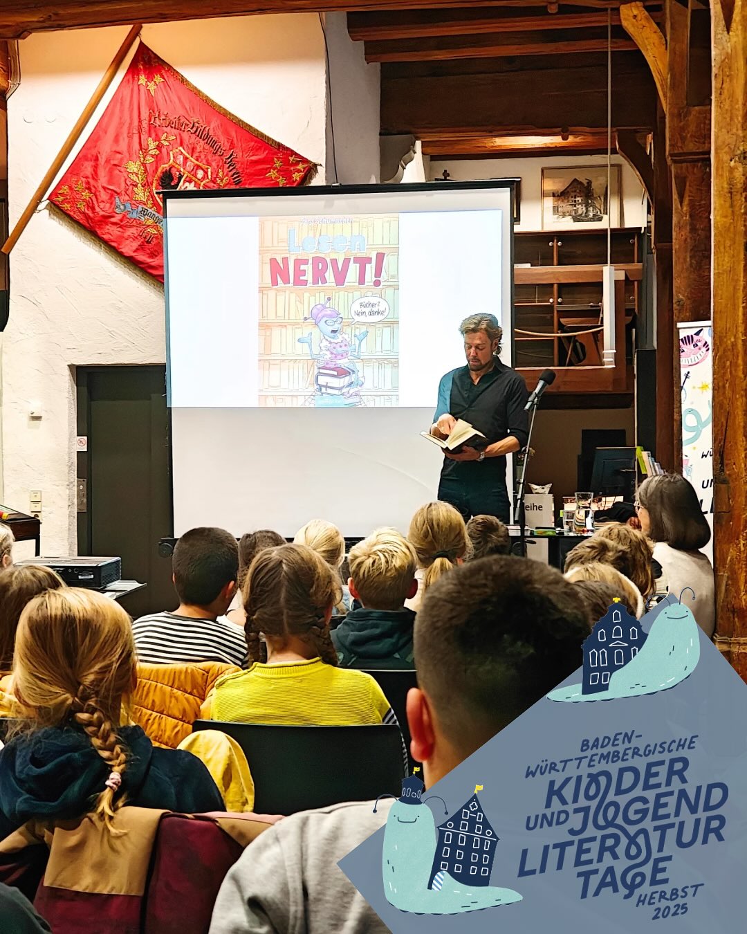 Dieser Alternativtext wurde KI-generiert: Eine Person steht vor einer großen Leinwand und liest ein Buch vor einer Gruppe von Menschen, die auf dem Boden sitzen. Im Hintergrund sind Dekorationen und ein Plakat mit Text zu sehen.