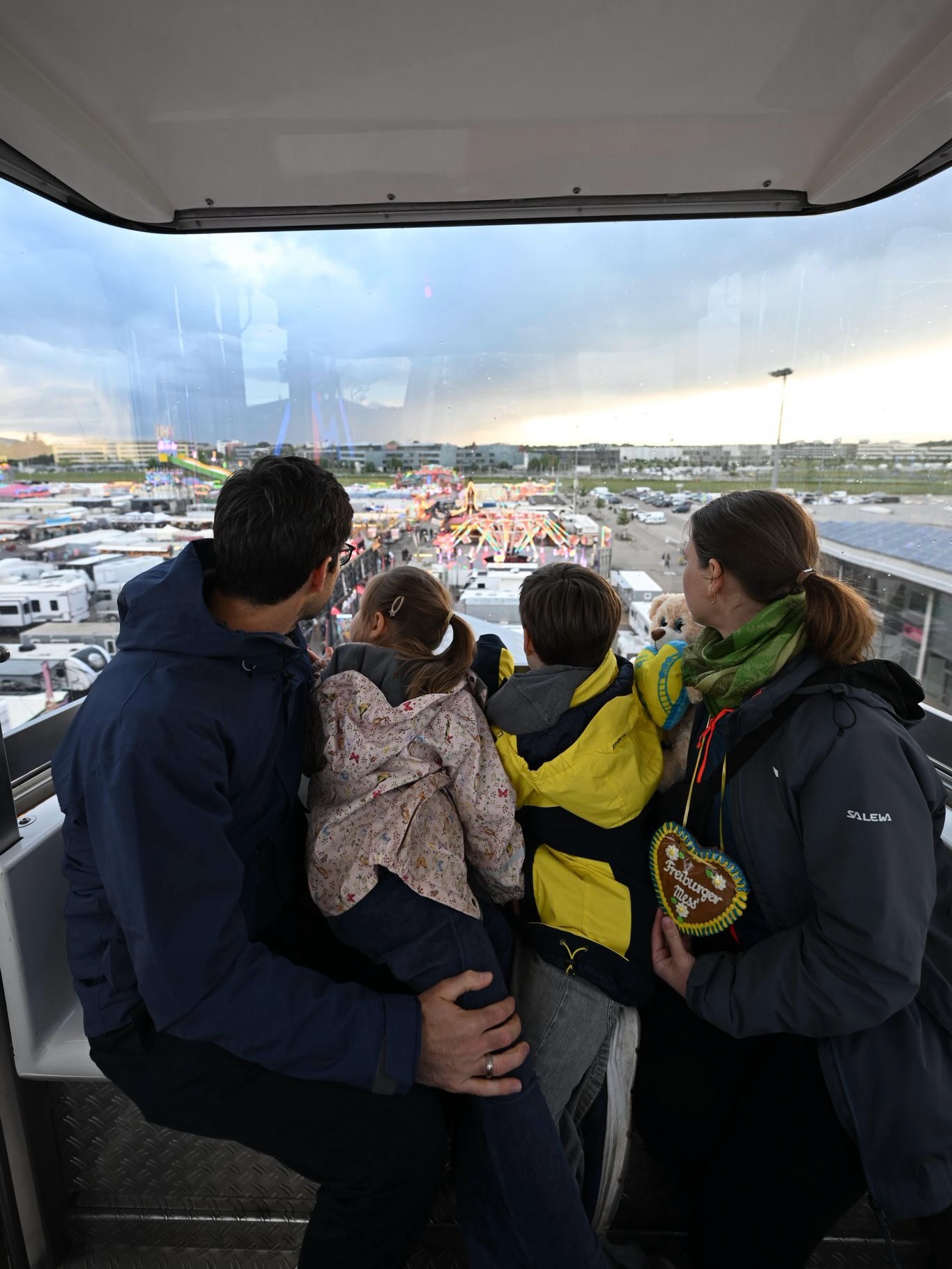 Eine Familie mit zwei Kindern blickt aus einer Fahrgastkabine auf ein belebtes Volksfest mit bunten Fahrgeschäften und Ständen. Eine Person hält ein herzförmiges Lebkuchenherz mit der Aufschrift „Freiburger Messe“. (KI-Alternativtext)