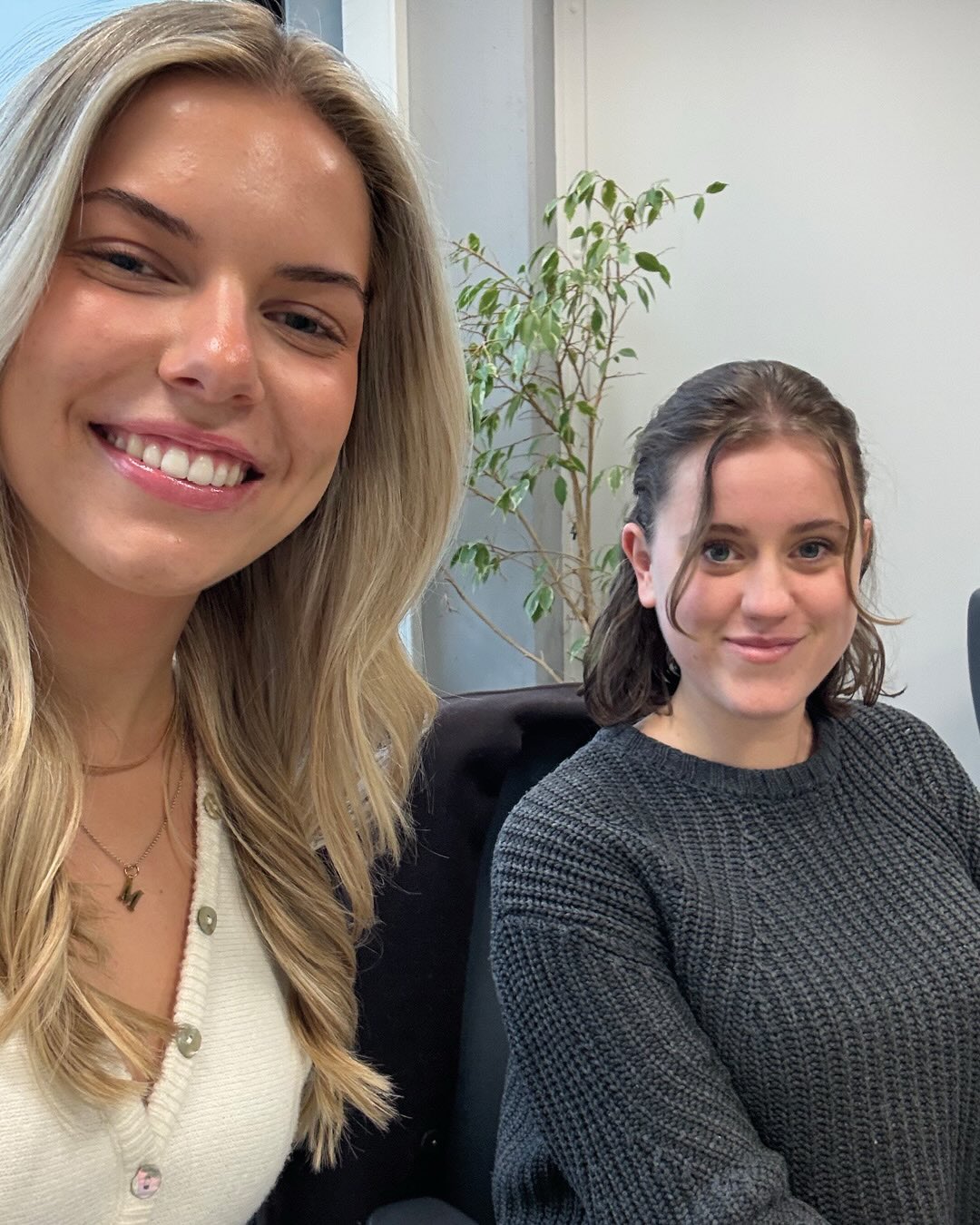 Dieser Alternativtext wurde KI-generiert: Zwei Personen lächeln in die Kamera. Im Hintergrund steht eine Pflanze und ein Bürostuhl.