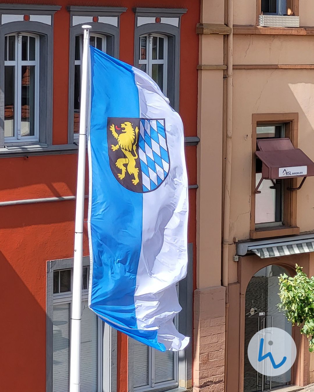 Dieser Alternativtext wurde KI-generiert: Eine blau-weiße Flagge weht vor einem Gebäude mit Fenstern und einem roten Anstrich. Auf der Flagge ist ein Wappen mit einem Löwen und Rauten abgebildet.