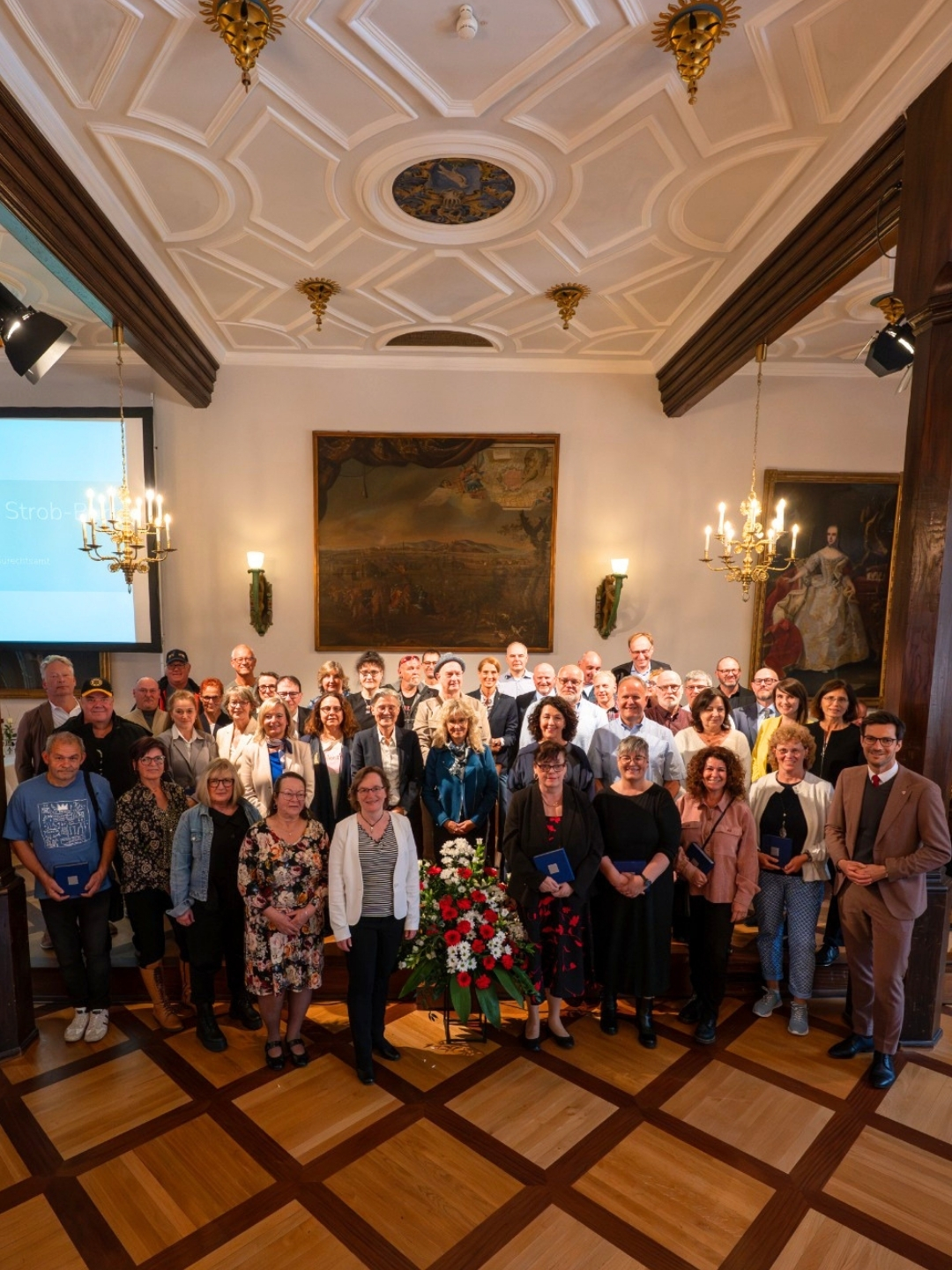 Gruppe von Menschen in einem repräsentativen Saal mit Stuckdecke, Kronleuchtern und Gemälden an den Wänden; einige halten blaue Urkunden, vorne steht ein Blumenstrauß mit roten und weißen Blüten. (KI-Alternativtext)