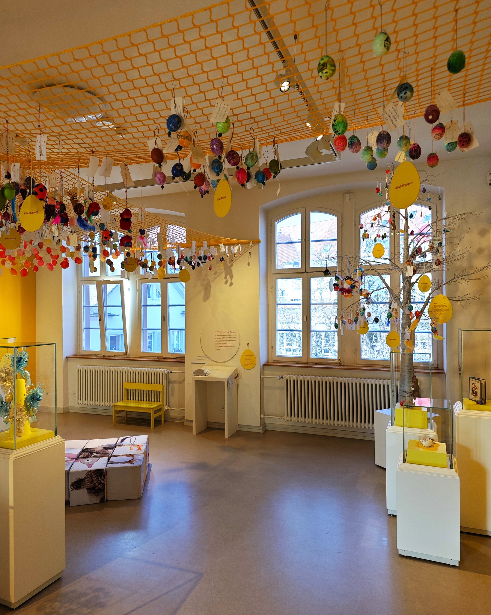 Dieser Alternativtext wurde KI-generiert: Ein Innenraum mit einer hellen Decke, von der viele bunte Objekte hängen. Im Raum sind Möbel und Fenster, sowie dekorative Elemente zu sehen.