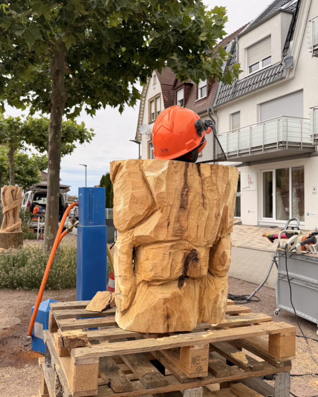 Holzskulptur mit grob behauener Oberfläche auf Paletten, darauf ein orangefarbener Schutzhelm mit der Aufschrift „RM“. Im Hintergrund Wohnhäuser, Bäume und Werkzeuge. (KI-Alternativtext)