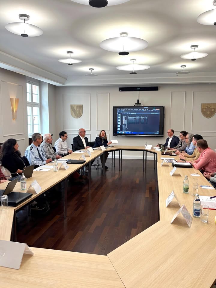 Dieser Alternativtext wurde KI-generiert: Ein langer Konferenztisch steht in einem hellen Raum mit mehreren Personen, die sich einem Bildschirm zuwenden. An den Wänden befinden sich Wappen und Deckenleuchten.