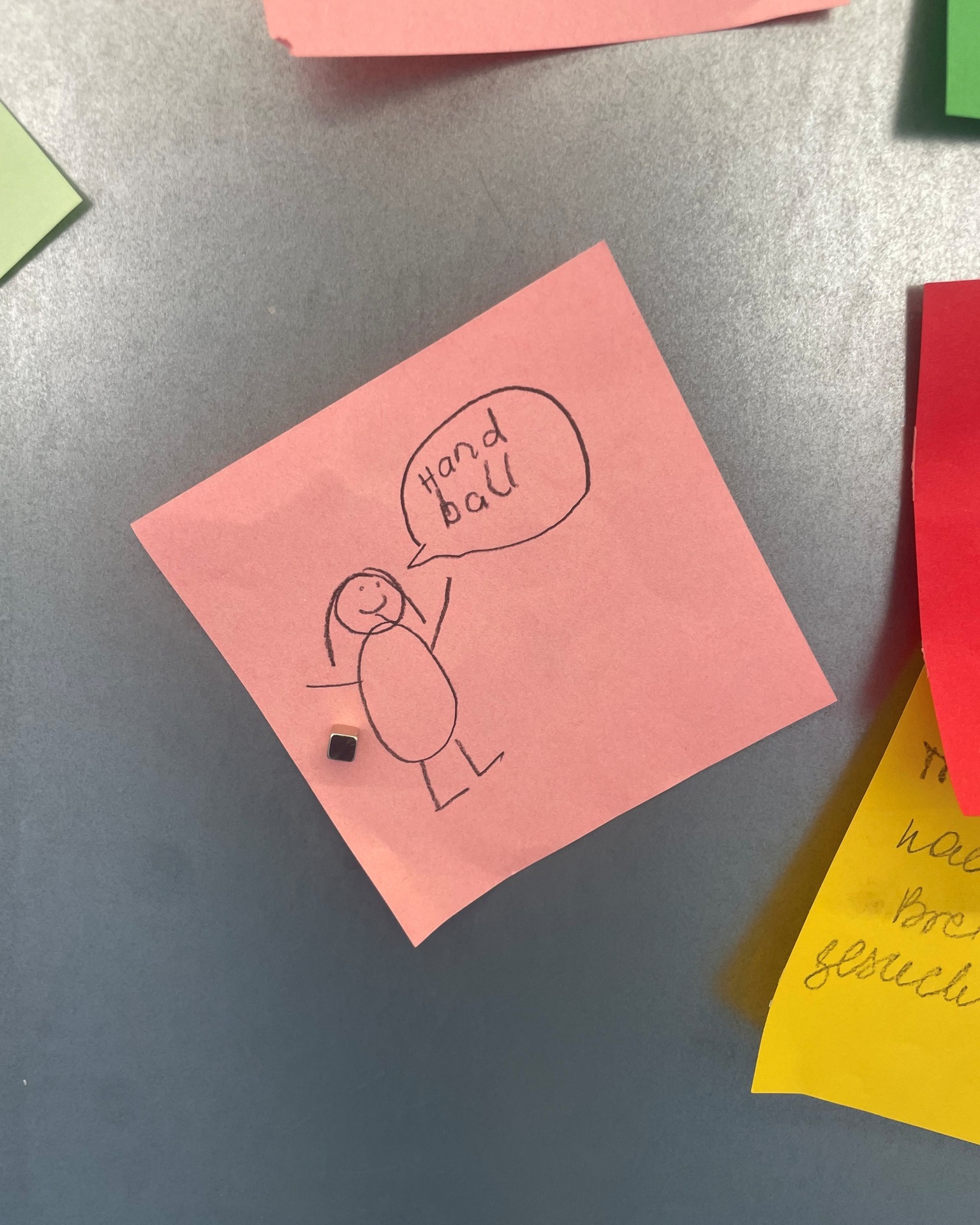 Ein rosa Haftnotiz mit einer Strichmännchen-Zeichnung und einer Sprechblase mit dem Text „Handball“ ist mit einem kleinen Magneten an einer grauen Oberfläche befestigt. Um die Notiz herum sind Teile weiterer bunter Haftnotizen zu sehen. (KI-Alternativtext)