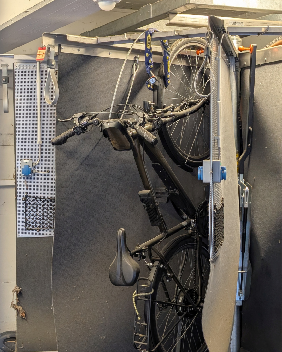 Ein schwarzes Fahrrad hängt an einer Wandhalterung in einem Raum mit dunkler Wand und Metallregalen. An der Halterung sind ein blauer Schlossgurt und ein kleines Netz befestigt. Links ist eine perforierte Metallplatte mit Haken und einem blauen Stecker zu sehen. (KI-Alternativtext)