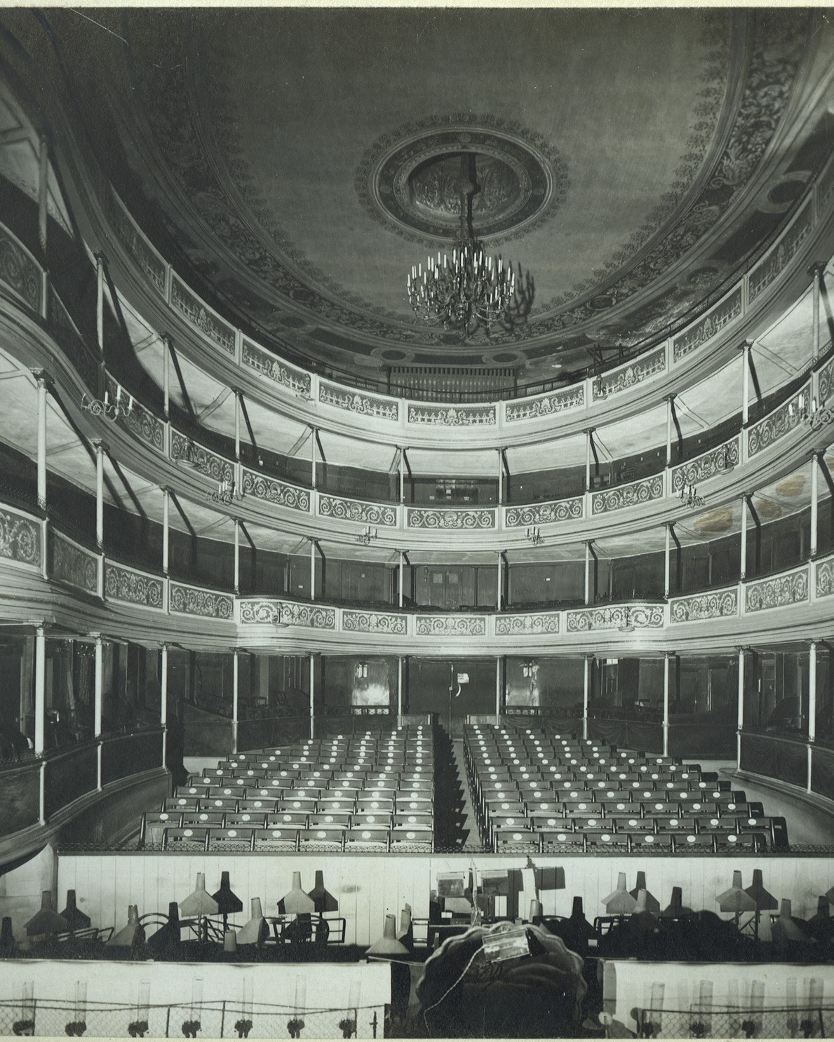 Dieser Alternativtext wurde KI-generiert: Schwarz-weißes Foto einer Innenansicht eines Theaters mit mehreren Sitzreihen und einer verzierten Decke mit einem Kronleuchter. Der Raum wirkt leer und weitläufig.