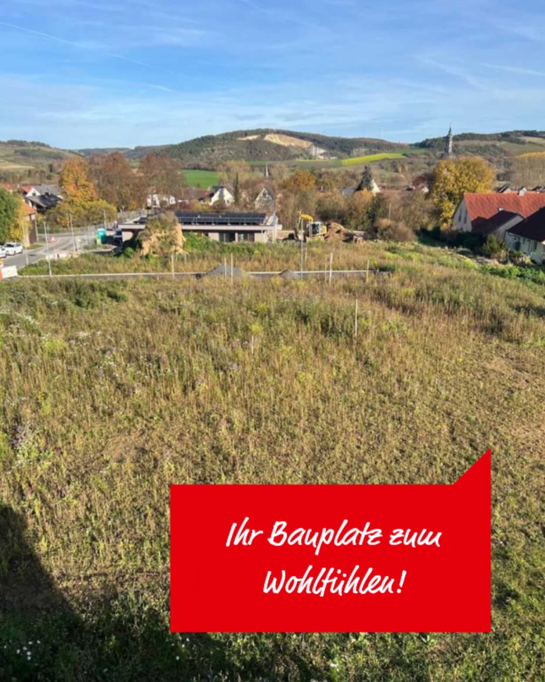 Dieser Alternativtext wurde KI-generiert: Ein hügeliges Landschaftsbild mit Häusern und einem großen Feld im Vordergrund. Auf dem Bild befindet sich roter Text, der auf einen Baugrundstück hinweist.