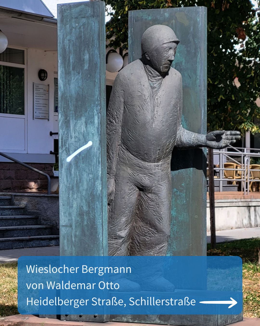 Dieser Alternativtext wurde KI-generiert: Eine metallene Figur steht vor einer Fassade und einigen Büschen. Unterhalb der Figur befindet sich ein blaues Schild mit einer weißen Pfeilzeichnung.