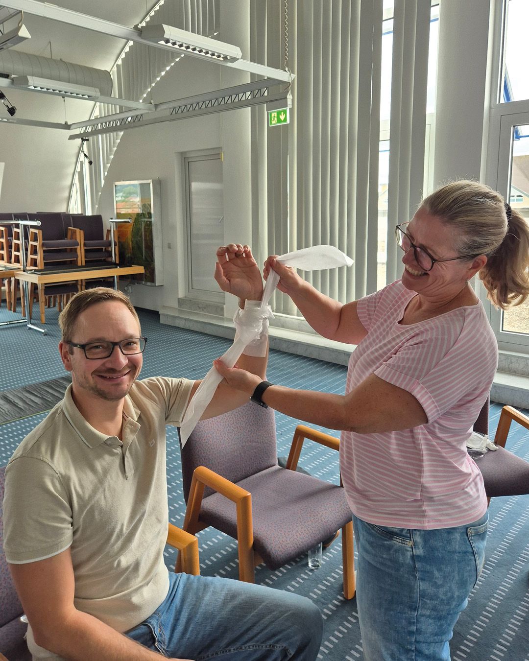 Eine stehende Person wickelt einen weißen Verband um den Unterarm einer sitzenden Person, während beide lächeln. Der Raum im Hintergrund enthält Stühle, Tische und große Fenster. (KI-Alternativtext)