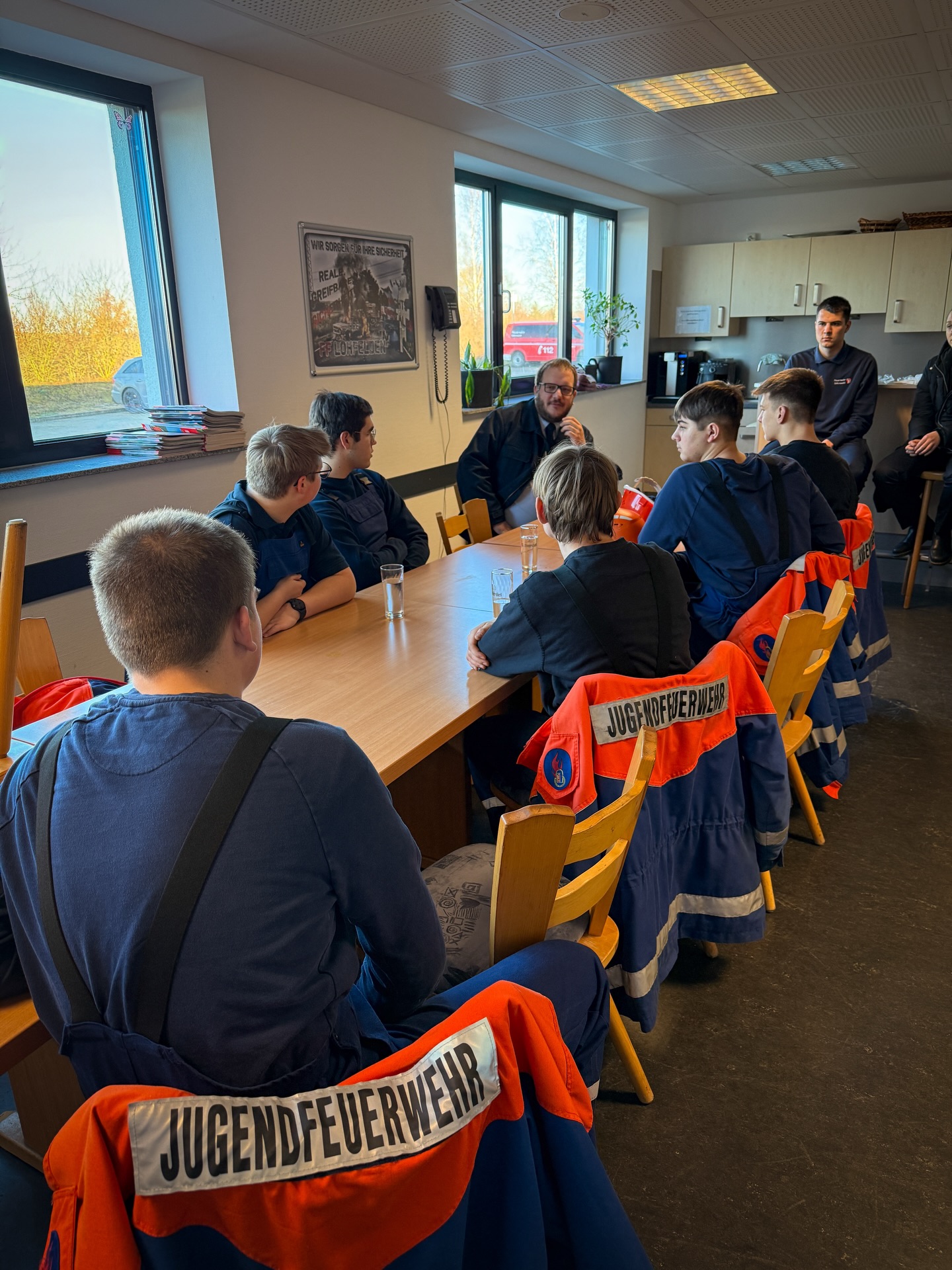 Mehrere Jugendliche in blauen Overalls sitzen um einen langen Holztisch in einem Raum. Auf den Stühlen liegen orangefarbene Jacken mit der Aufschrift „JUGENDFEUERWEHR“. Am Kopfende des Tisches sitzt eine erwachsene Person, während an der Wand ein Poster mit der Aufschrift „FF LOHN FELDEN“ hängt. (KI-Alternativtext)