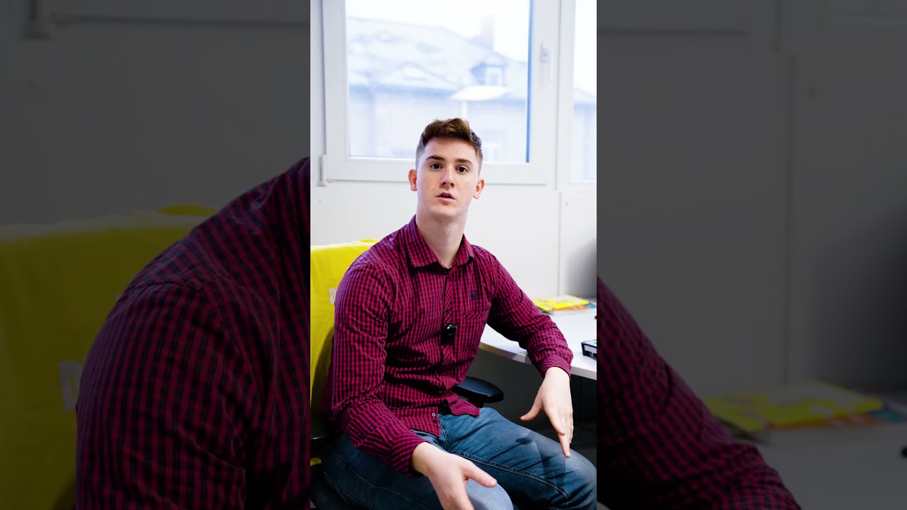 Dieser Alternativtext wurde KI-generiert: Eine Person sitzt auf einem gelben Stuhl und blickt zur Seite. Im Hintergrund ist ein Fenster und ein Schreibtisch zu sehen.