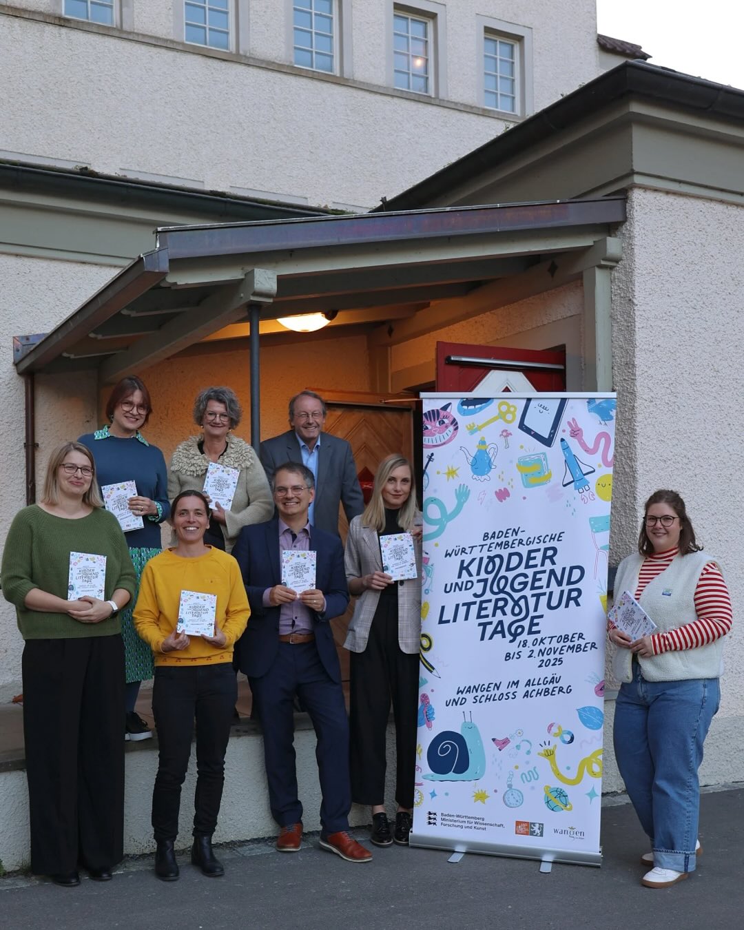 Dieser Alternativtext wurde KI-generiert: Eine Gruppe von Menschen steht vor einer hellen Fassade mit einem Plakat. Das Plakat bewirbt Kindertage und Literaturtage in Wangen im Allgäu und Schloss Achberg.