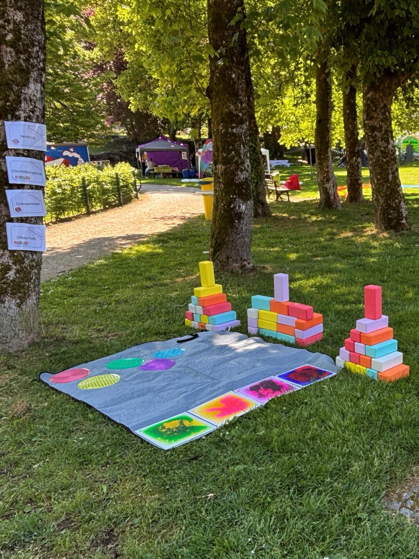 Dieser Alternativtext wurde KI-generiert: Ein farbenfrohes Bild zeigt eine Grünfläche mit mehreren Personen und bunten Bauklötzen im Vordergrund. Im Hintergrund sind weitere Menschen und Verkaufsstände zu erkennen.