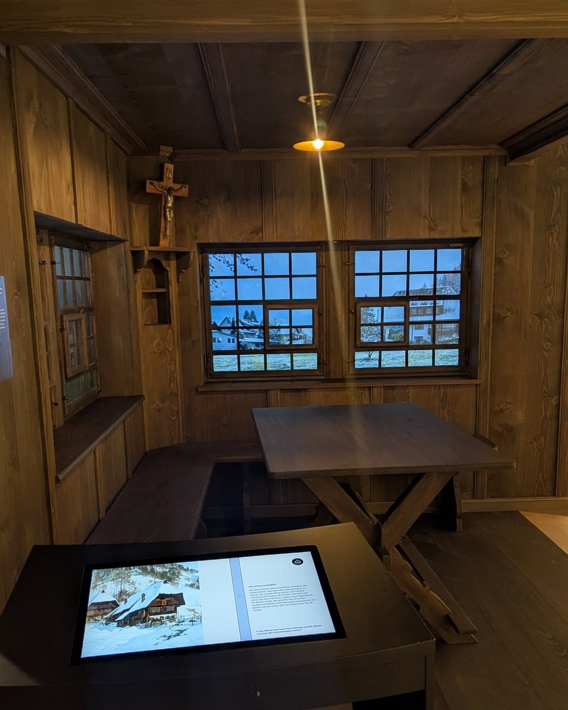 Dieser Alternativtext wurde KI-generiert: Ein Innenraum mit Holzvertäfelung und einem Fenster, das einen Blick auf eine verschneite Landschaft bietet. Im Raum befinden sich ein Tisch, Bänke und ein Bildschirm mit einem Bild.