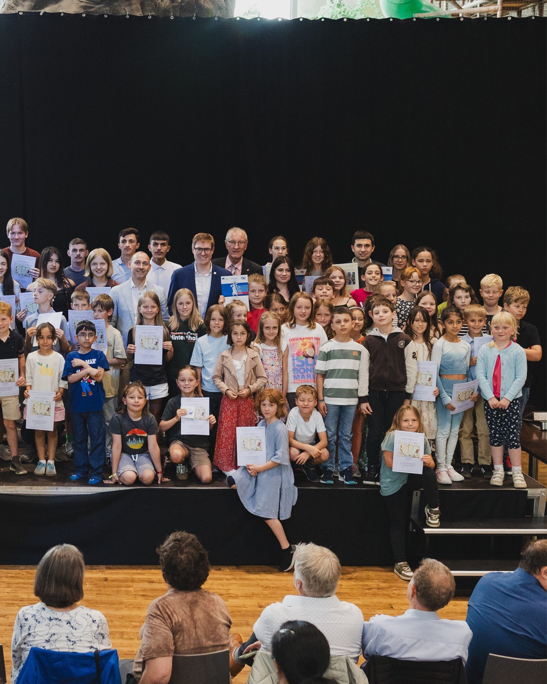 Gruppe von Kindern und Erwachsenen auf einer Bühne, die Urkunden halten und in Richtung Publikum blicken. Im Vordergrund sitzen Zuschauer auf Stühlen. (KI-Alternativtext)