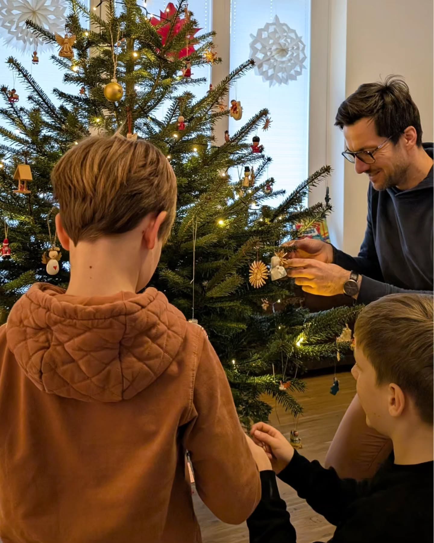 Dieser Alternativtext wurde KI-generiert: Ein geschmückter Weihnachtsbaum steht im Raum. Mehrere Personen sind dabei, den Baum gemeinsam zu dekorieren.