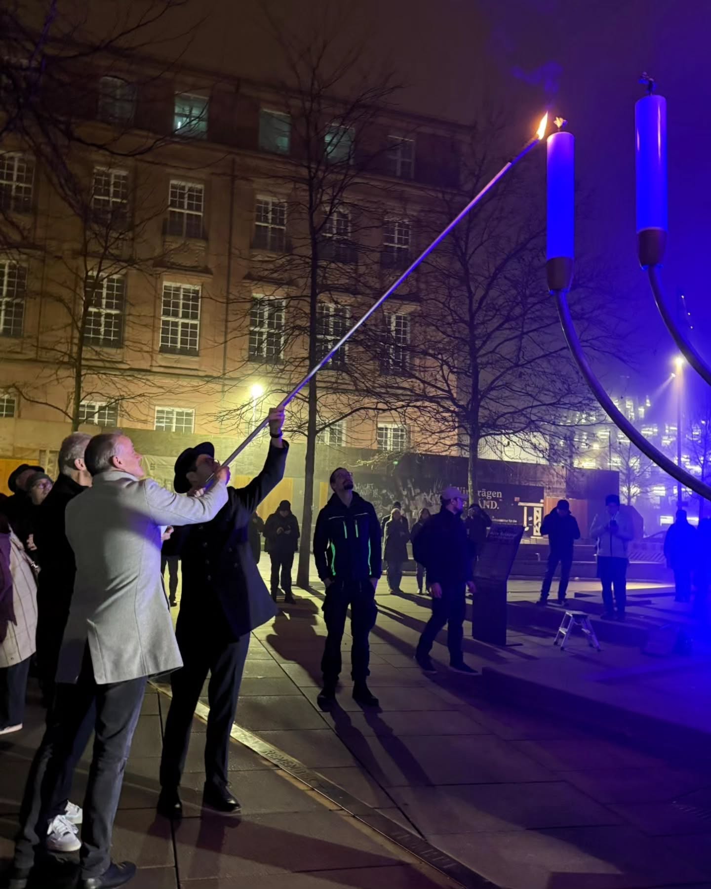 Dieser Alternativtext wurde KI-generiert: Ein Schwarz-Weiß-Foto zeigt eine nächtliche Szene mit einer beleuchteten Installation, die in die Höhe gehalten wird. Im Hintergrund sind Gebäude und Menschen zu erkennen.