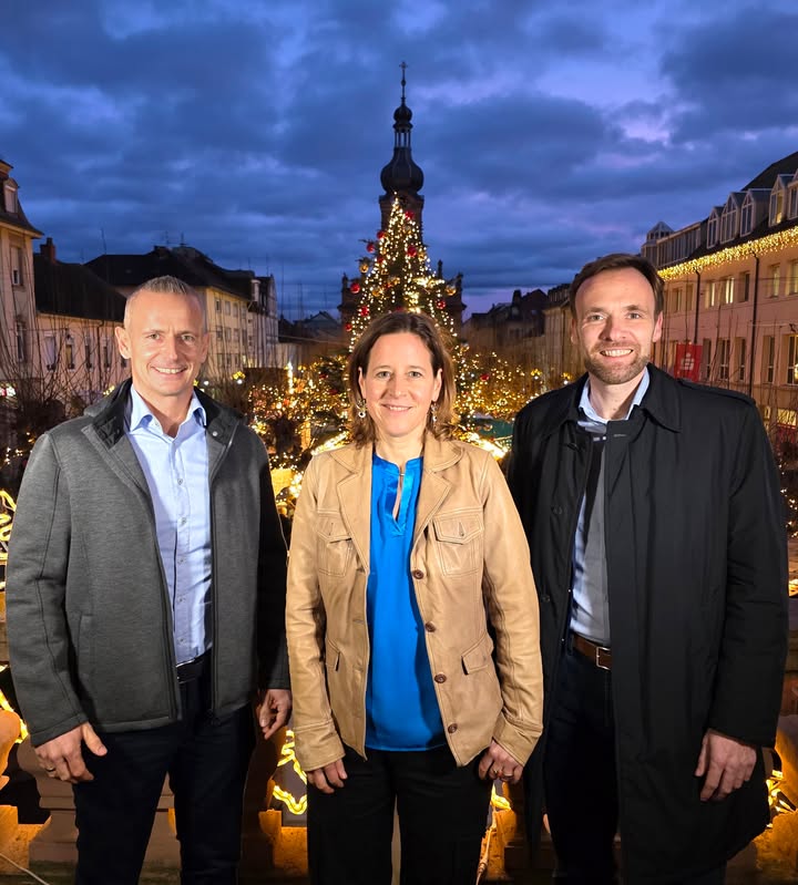 Dieser Alternativtext wurde KI-generiert: Drei Personen stehen vor einem beleuchteten Weihnachtsmarkt in der Dämmerung. Im Hintergrund ist ein hoher Turm mit Weihnachtsdekorationen zu sehen.