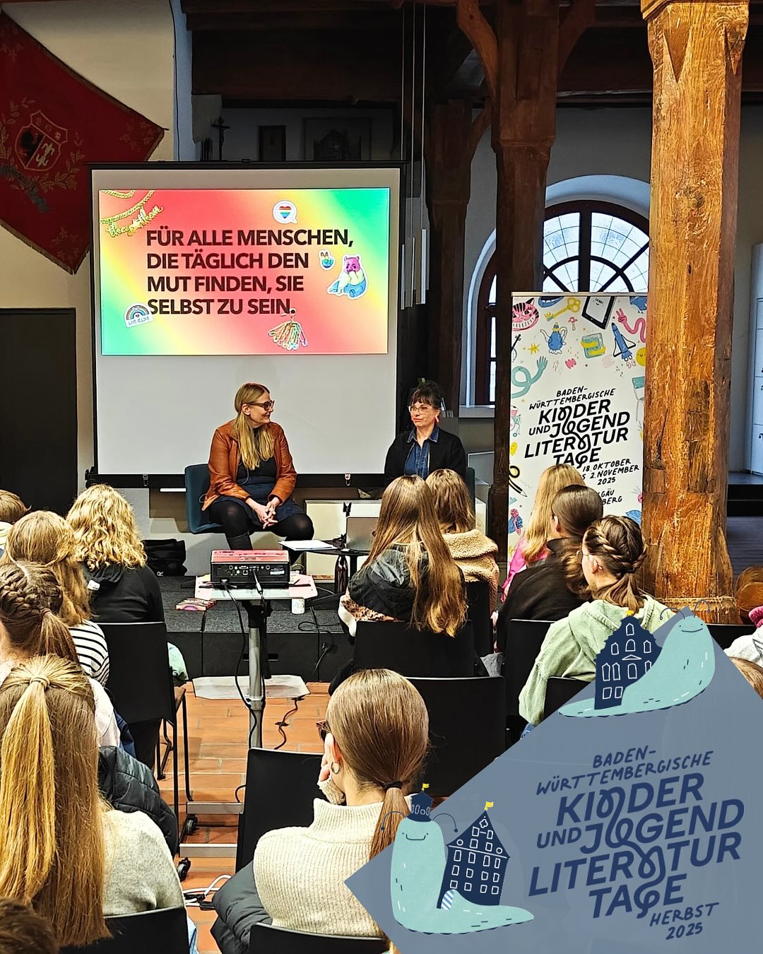 Dieser Alternativtext wurde KI-generiert: Schwarz-weißes Foto einer Person, die auf einem Stuhl sitzt und sich mit einer Gruppe von Personen unterhält. Im Hintergrund befinden sich Plakate und Fenster.