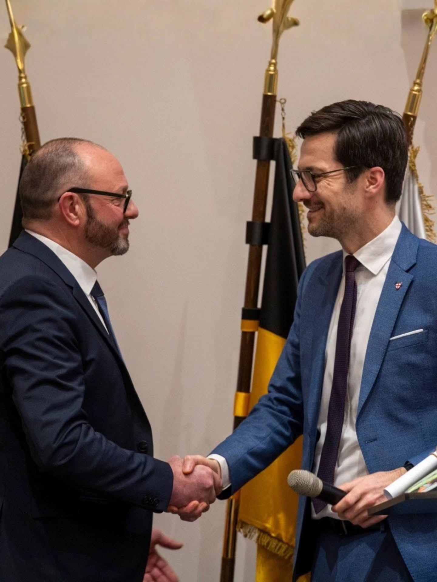 Dieser Alternativtext wurde KI-generiert: Zwei Personen in Anzügen geben sich die Hand. Im Hintergrund sind Fahnen und ein Mikrofon zu sehen.