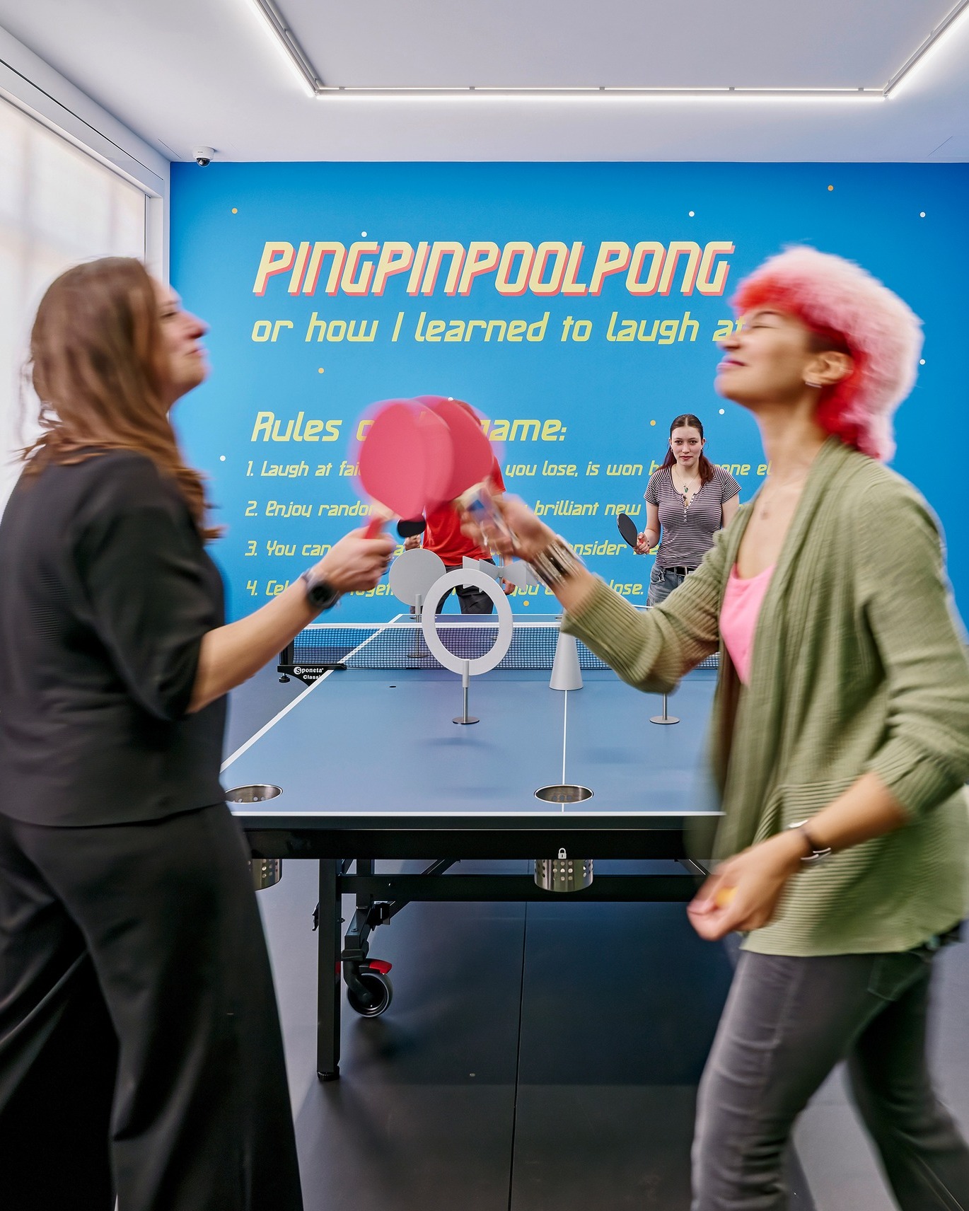 Zwei Personen spielen mit rosa Schlägern ein Spiel namens 'PINGPINPOOLPONG' an einem blauen Tisch mit Löchern und Hindernissen. Im Hintergrund steht an einer blauen Wand der Titel sowie Spielregeln, während weitere Personen zuschauen. Die Person links trägt dunkle Kleidung, die Person rechts hat rosa Haare und eine grüne Jacke. (KI-Alternativtext)
