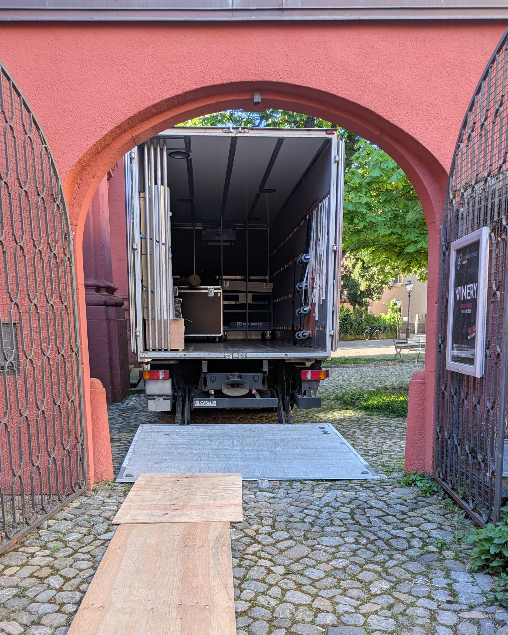 Dieser Alternativtext wurde KI-generiert: Ein offener Lastwagen steht unter einem steinernen Torbogen. Im Hintergrund sind Gebäude und Bäume zu sehen.