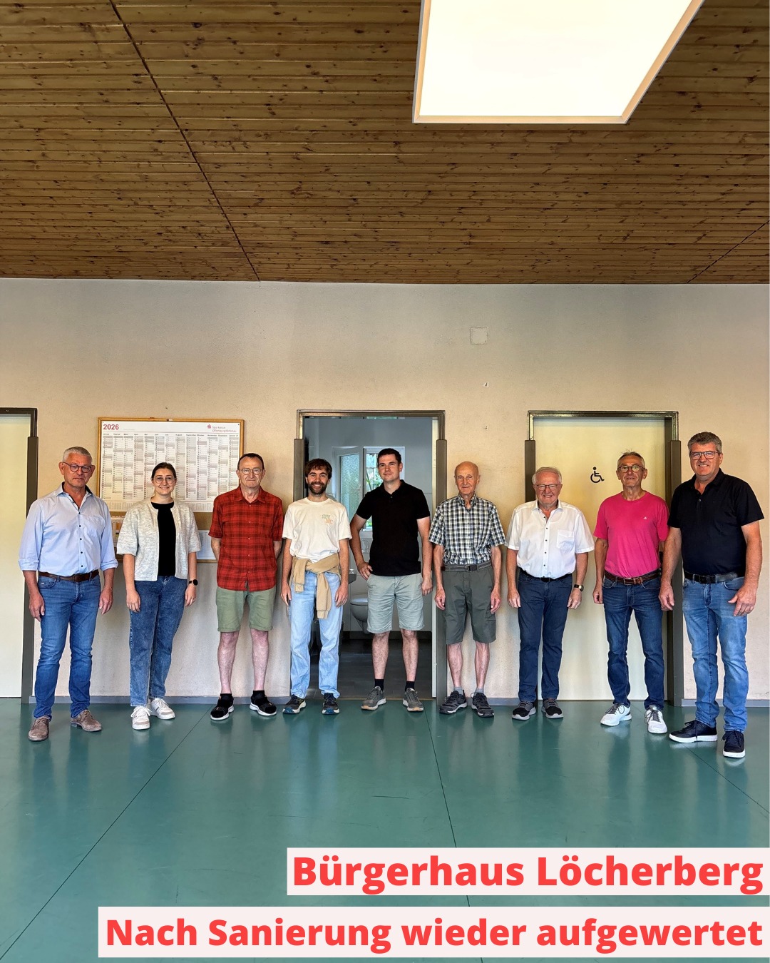 Neun Personen stehen nebeneinander in einem Raum mit Holzdecke und grünem Boden. Im Hintergrund sind Türen und ein Wandkalender zu sehen. Ein Textfeld unten lautet: "Bürgerhaus Löcherberg Nach Sanierung wieder aufgewertet". (KI-Alternativtext)