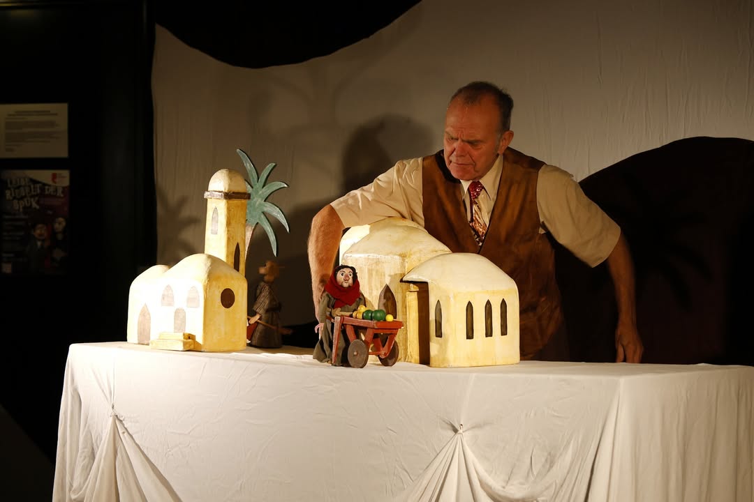 Dieser Alternativtext wurde KI-generiert: Ein Mann steht vor einem Tisch mit einer kleinen Krippendarstellung. Die Szene ist in warmen Farbtönen gehalten und wirkt wie eine Puppenvorführung.