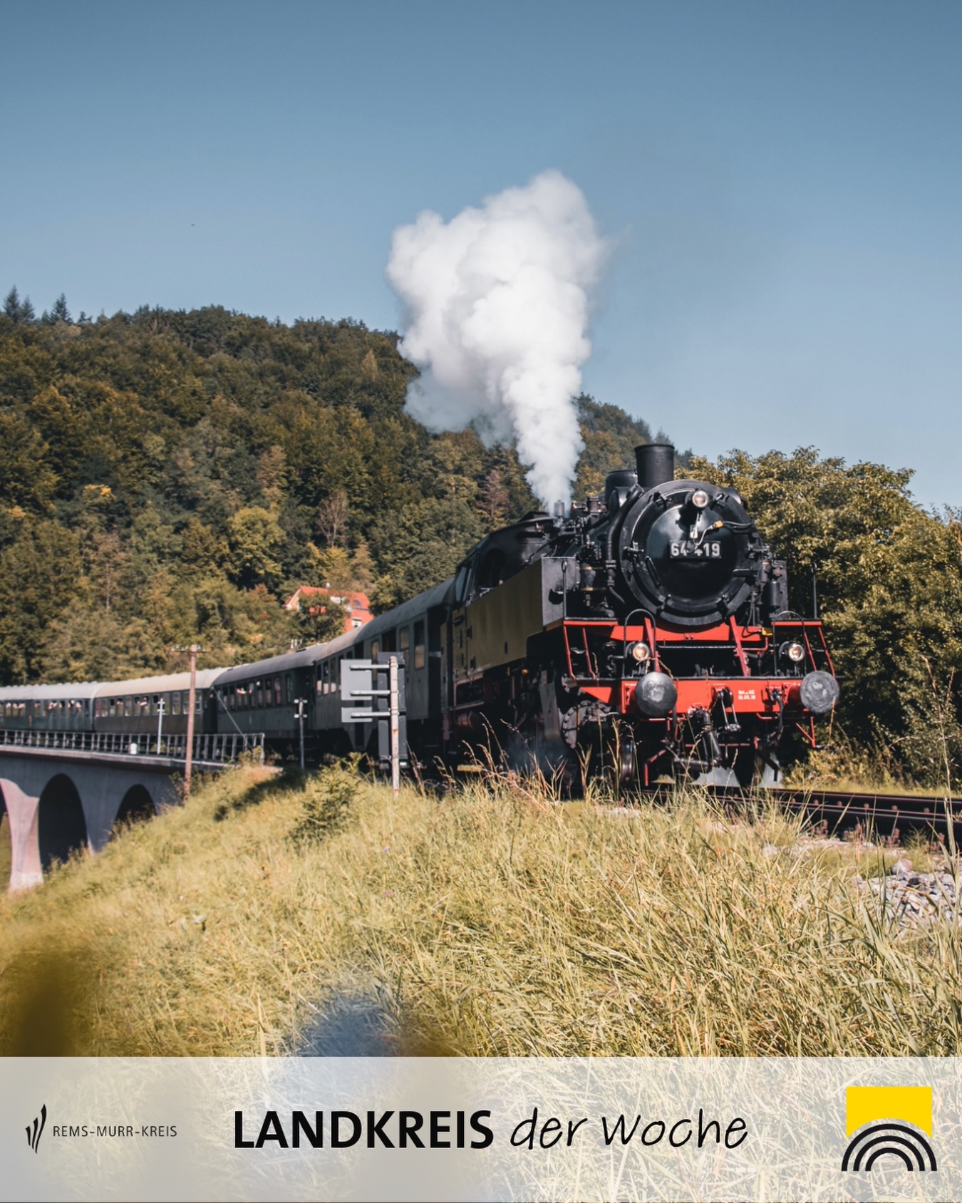 Dieser Alternativtext wurde KI-generiert: Eine Dampflokomotive fährt auf einer Strecke über eine Brücke vor einer bewaldeten Hügellandschaft. Das Bild ist farbig und zeigt eine idyllische Szene.