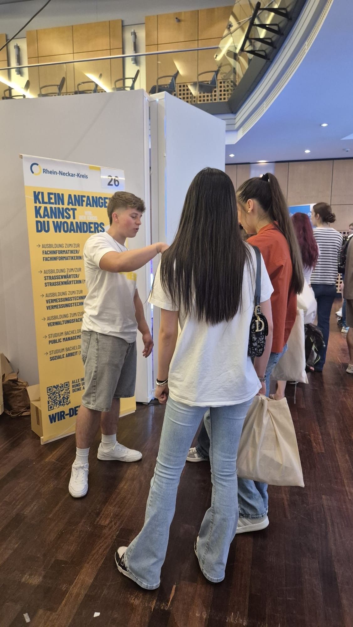 Eine Person in weißem T-Shirt und Shorts spricht vor einem gelben Standbanner mit zwei anderen Personen. Das Banner trägt die Aufschrift „Rhein-Neckar-Kreis“ und „KLEIN ANFANGEN KANNST DU WOANDERS“ sowie eine Liste von Ausbildungsberufen. Im Hintergrund stehen weitere Personen in einer modernen Halle. (KI-Alternativtext)