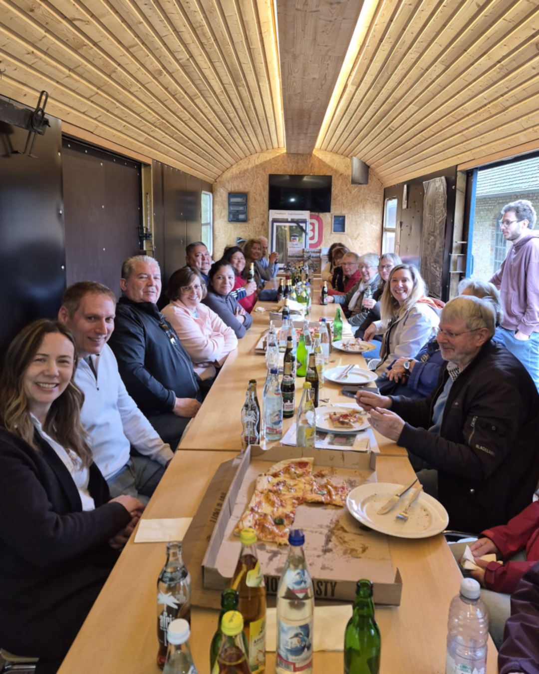 Mehrere Personen sitzen an einem langen Holztisch in einem Raum mit Holzdecke und essen Pizza, trinken Getränke aus Flaschen und schauen in die Kamera. Einige lächeln, eine Person steht am Ende des Tisches. (KI-Alternativtext)