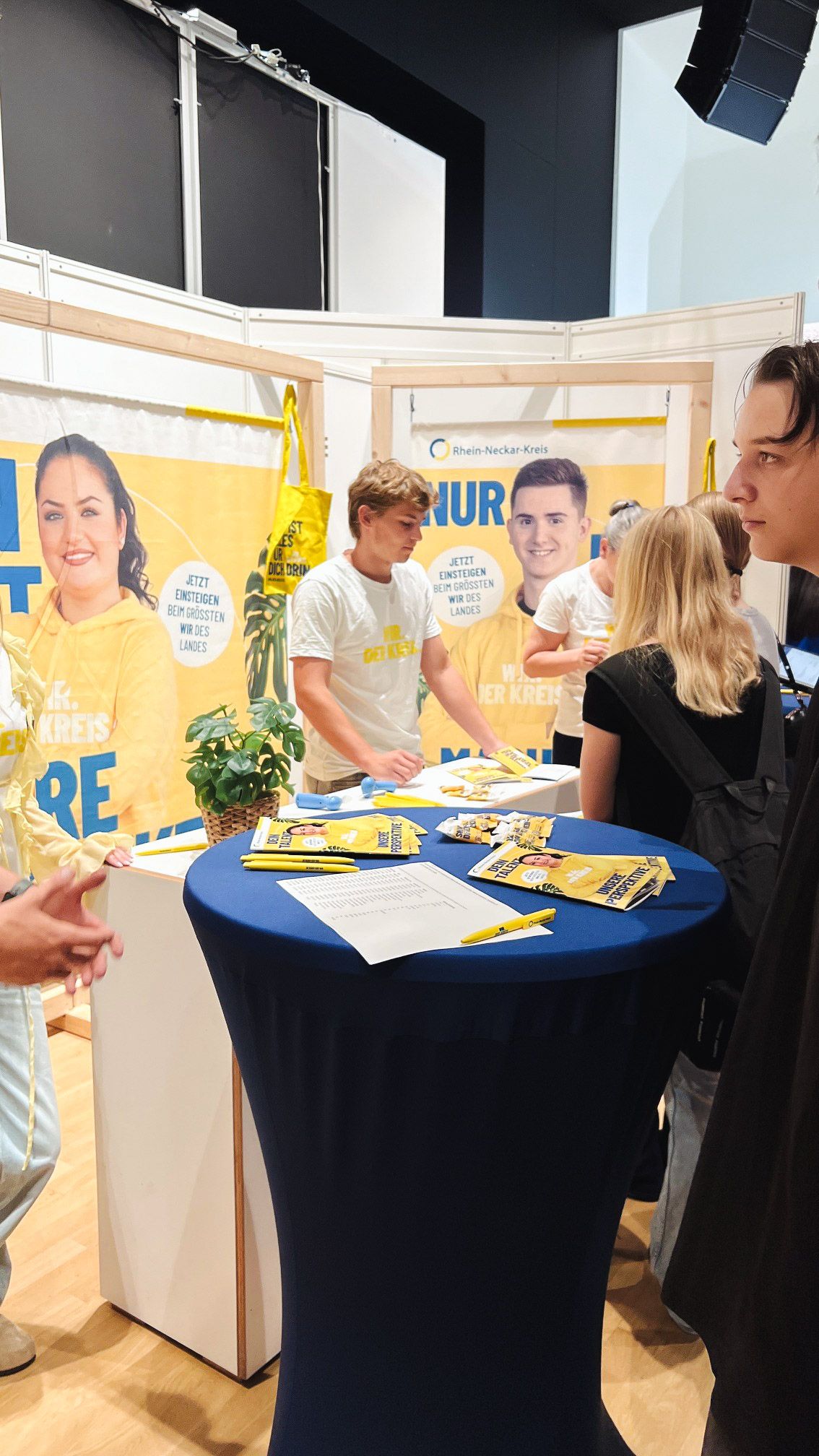 Ein Messestand des Rhein-Neckar-Kreises mit einem blauen Hochtisch, auf dem gelbe Flyer, Stifte und ein Dokument liegen. Mehrere Personen stehen am Stand, wobei eine Person hinter dem Tresen steht und andere davor. Im Hintergrund hängen große Banner mit dem Slogan "Jetzt einsteigen beim größten Wir des Landes" und Abbildungen von Personen in gelben Hoodies. (KI-Alternativtext)