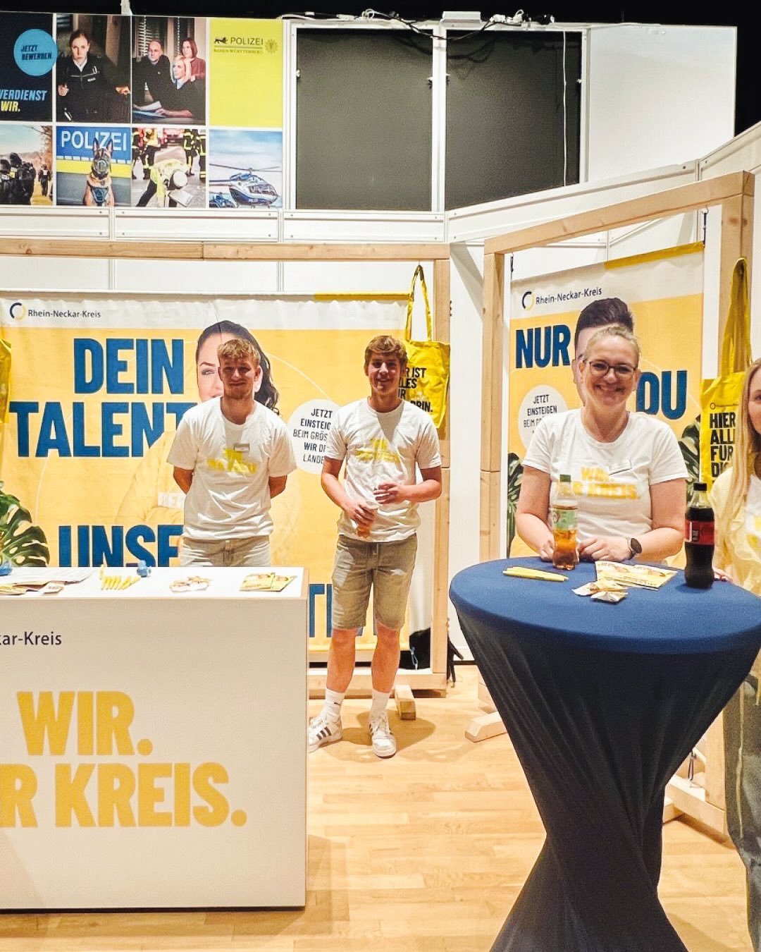 Messestand des Rhein-Neckar-Kreises mit vier Personen in weißen T-Shirts, die hinter einem weißen Tresen und einem blauen Hochtisch stehen. Im Hintergrund sind große gelbe Banner mit den Texten „Dein Talent“ und „Nur Du“ sowie ein Plakat der Polizei zu sehen, während auf dem Tresen der Slogan „Wir. Unser Kreis.“ steht. (KI-Alternativtext)