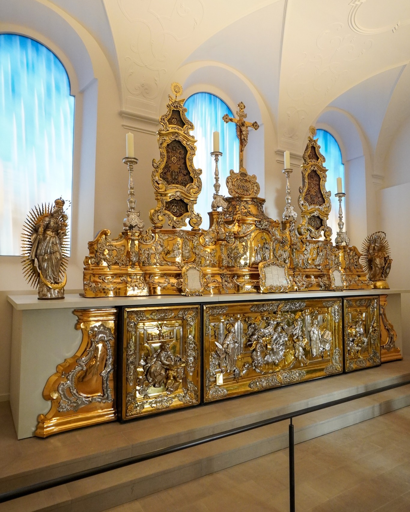Dieser Alternativtext wurde KI-generiert: Eine kunstvolle barocke Anordnung steht auf einer verzierten Kommode. Die Dekoration besteht aus goldenen Ornamenten und religiösen Figuren vor hohen, spitzbogenförmigen Fenstern.