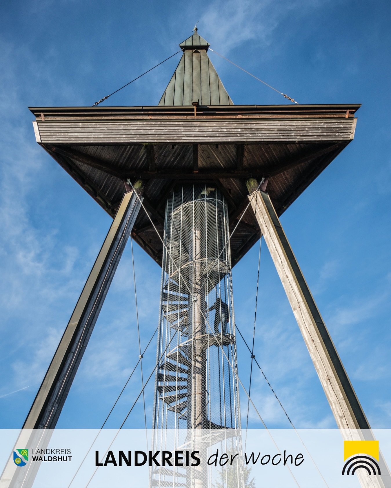 Ein hölzerner Aussichtsturm erhebt sich gegen einen blauen Himmel. Eine Wendeltreppe führt nach oben.