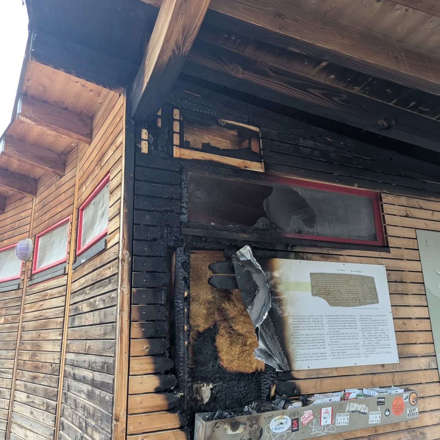 Dieser Alternativtext wurde KI-generiert: Ein Holzgebäude zeigt deutliche Brandschäden an der Fassade. Ein Informationsschild ist ebenfalls beschädigt und mit Brandspuren bedeckt.