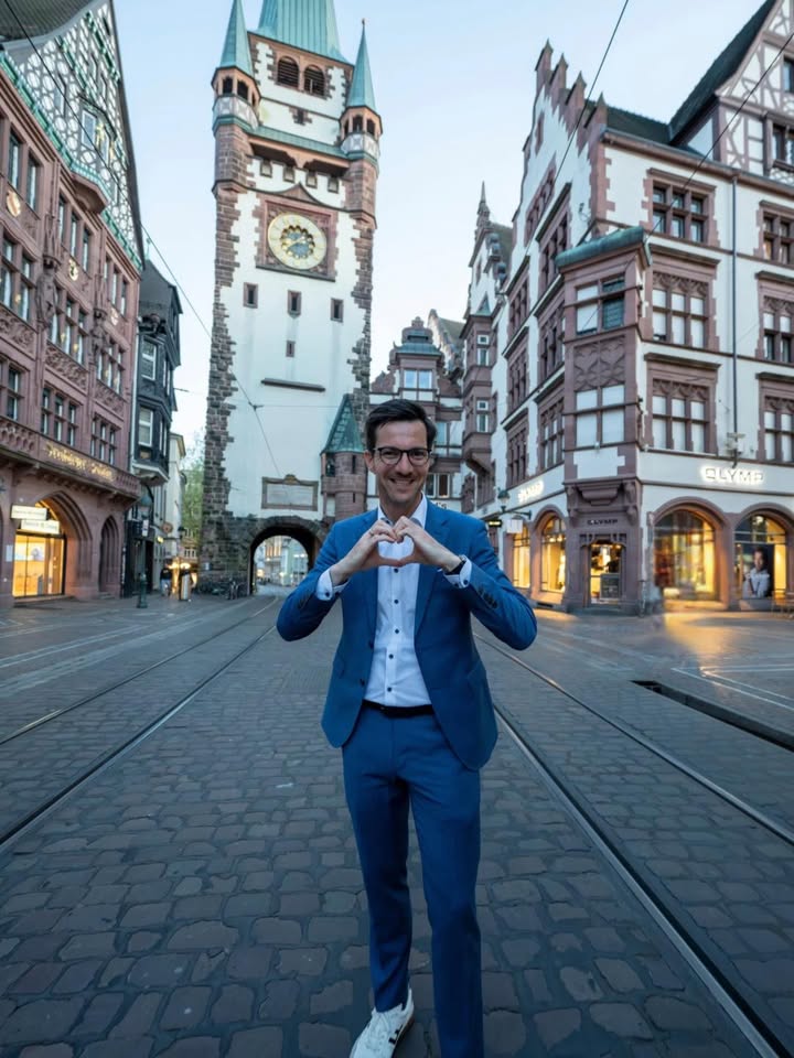 Dieser Alternativtext wurde KI-generiert: Ein historischer Straßenabschnitt mit Fachwerkhäusern und einem markanten Turm. Eine Person formt mit den Händen ein Herz vor der Kulisse.
