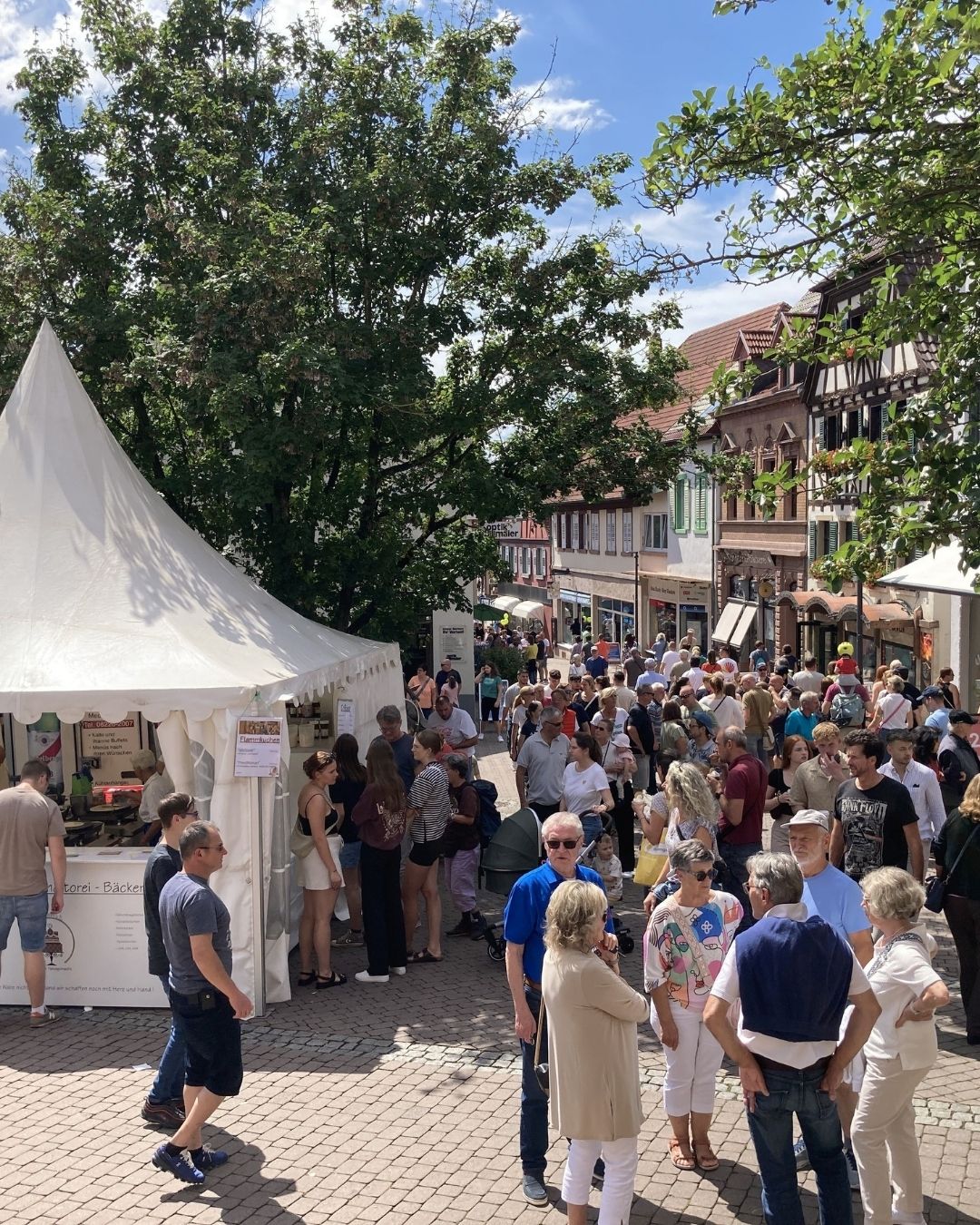 Dieser Alternativtext wurde KI-generiert: Eine belebte Stadtstraße mit vielen Menschen und kleinen Marktständen. Im Hintergrund sind traditionelle Fachwerkhäuser zu sehen.